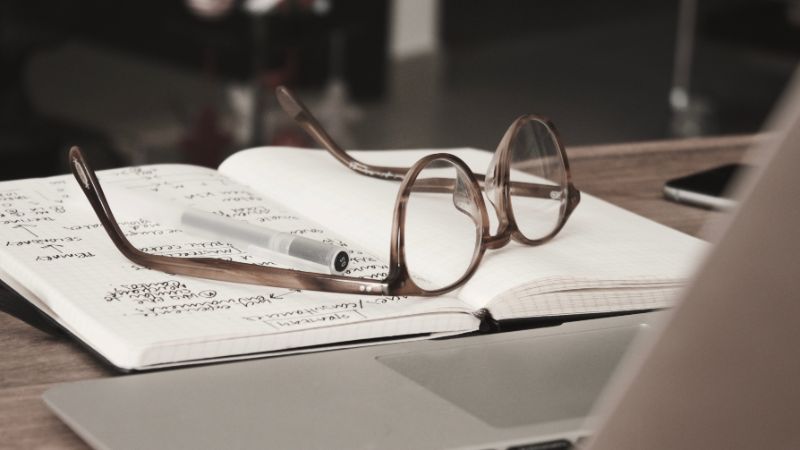 Hypnotherapist notepad and pen within writing and glasses on top, with a laptop partly visible.