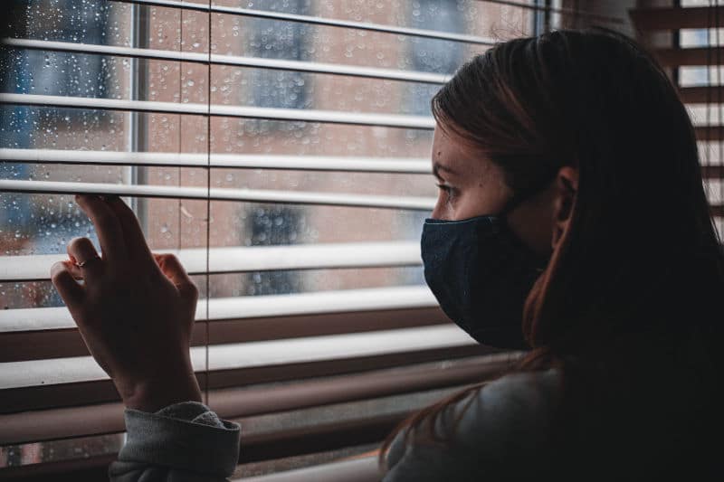 Anxiety and Woman in mask, in lockdown, staring sadly through a rainy window to the outside world